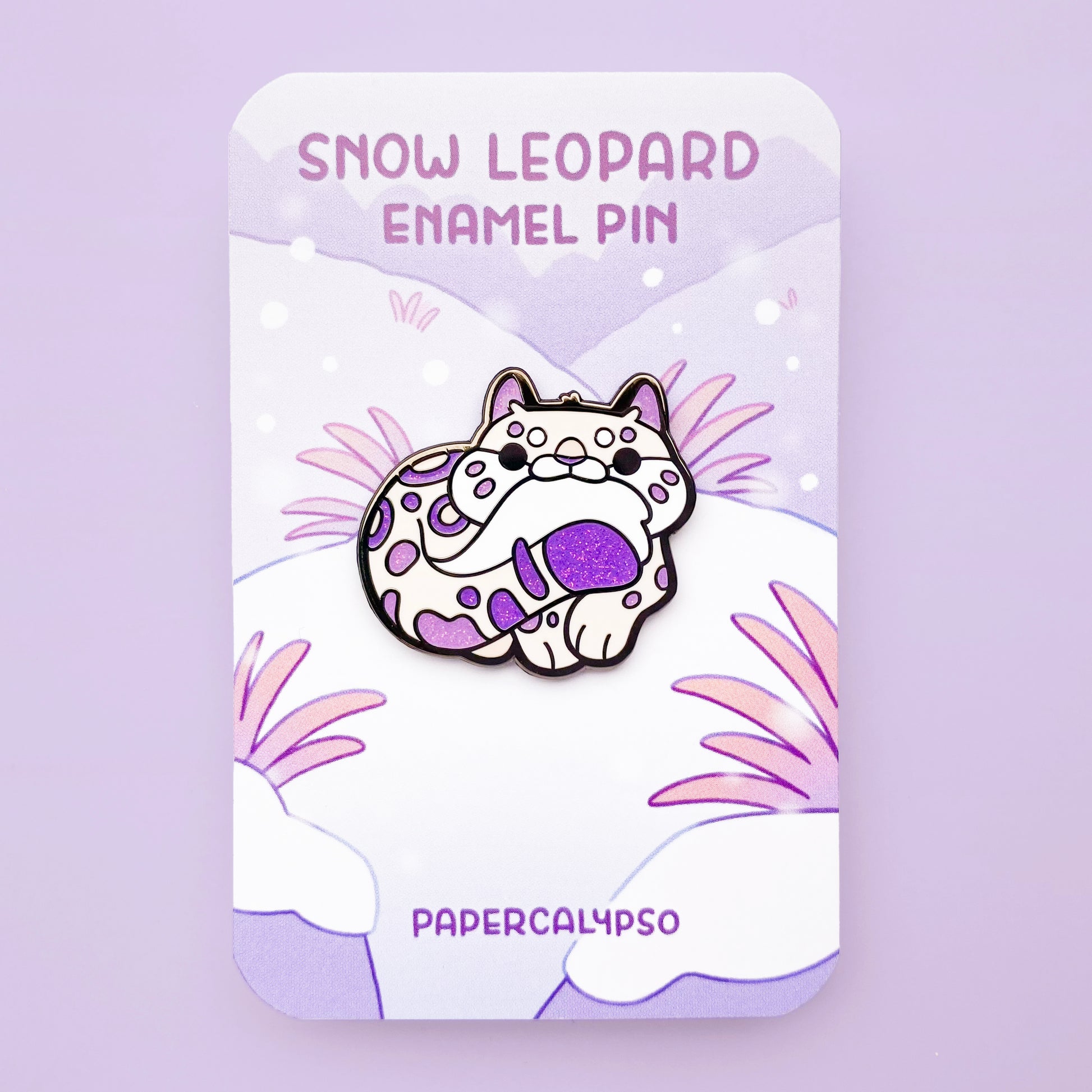 A black nickel hard enamel pin of a cute snow leopard biting its own tail. He's a cream colour with purple glitter spots.