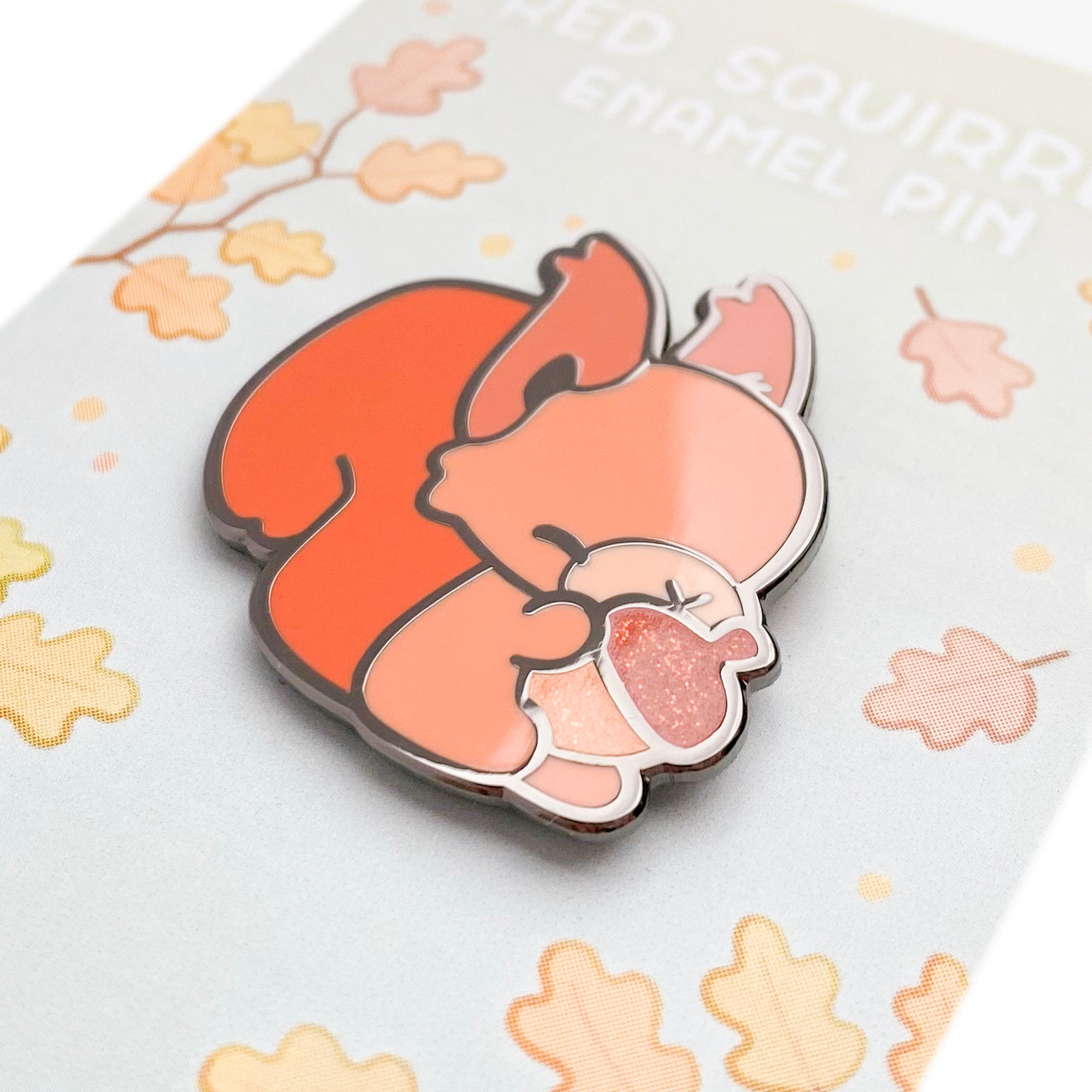 A close-up of the pin so you can see the details. The glitter accents sparkle in the light. The pin backing is an autumnal oak tree forest and the title reads "Red Squirrel Enamel Pin".