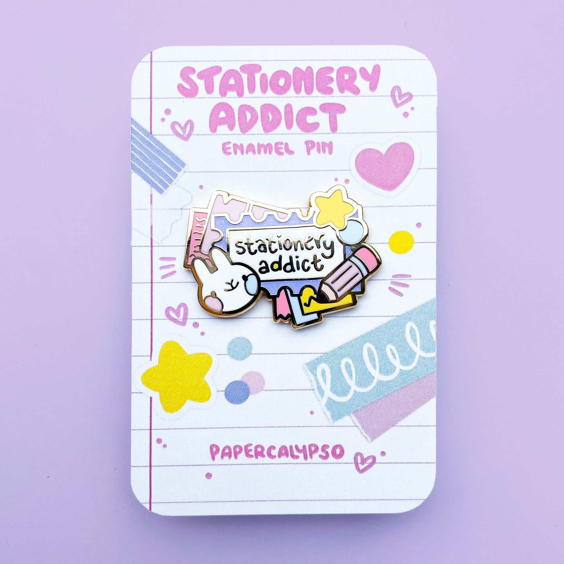 A gold hard enamel pin of a postage stamp with stationery around it. In the middle is the text "stationery addict". There is also a writing pencil, some star shaped stickers, a rabbit, and some tickets and papers. The stamp has purple glitter enamel. The colour scheme is pastel and has pinks, yellows and blues.