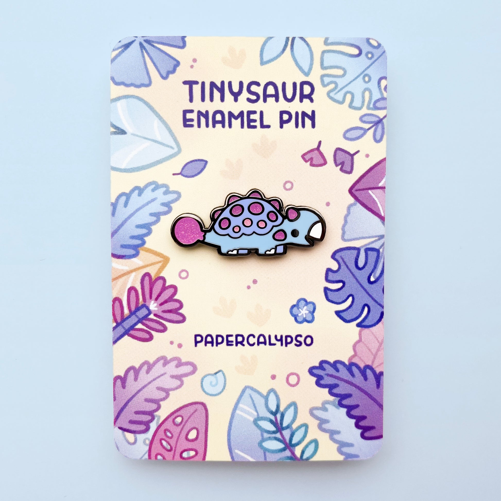 A black nickel hard enamel pin of a pink and blue dinosaur ankylosaurus. He has glittery spikes and a big smile.