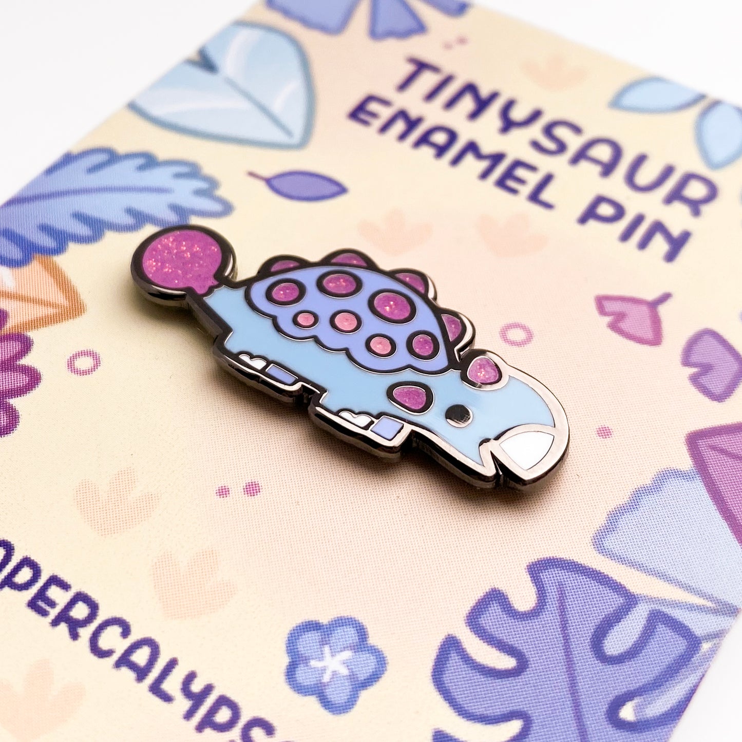 A close-up of the pin so you can see the details. The glitter accents sparkle in the light. The pin backing is a colorful prehistoric jungle scene with the title "Tinysaur Enamel Pin".