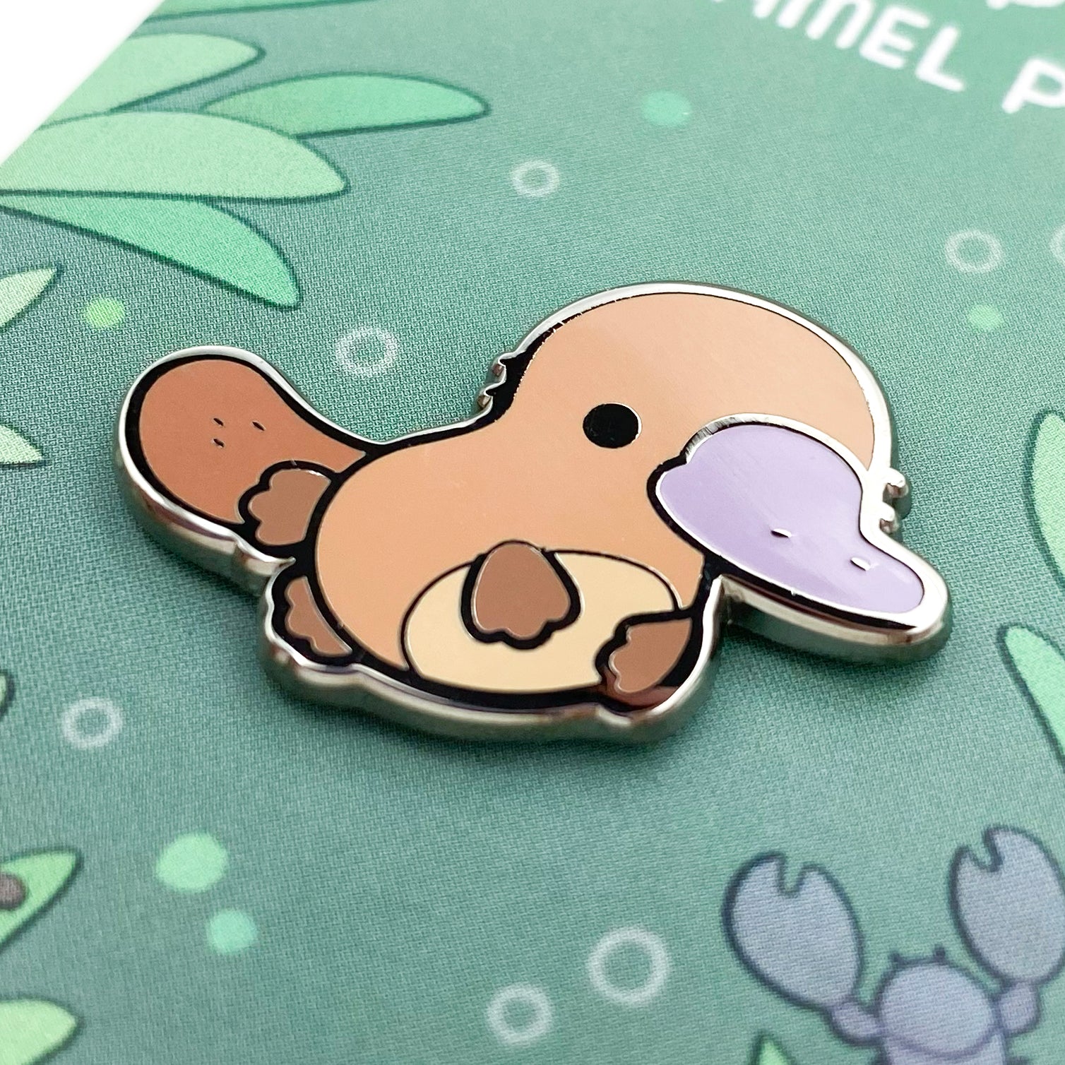 A close-up of the pin. The pin backing is an underwater scene with australian plants, a yabby and a waterbug. It has the title "Platypus Enamel Pin."