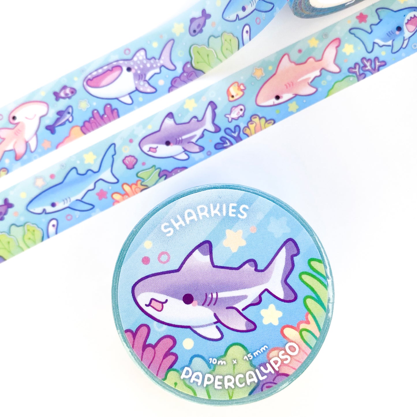 the roll of washi tape is packaged with shrink wrap plastic and a round sticker on one side. On it is an adorable happy shark surrounded by corals and stars. It reads "Sharkies, 10 metres by 15 milimetres, by Papercalypso"