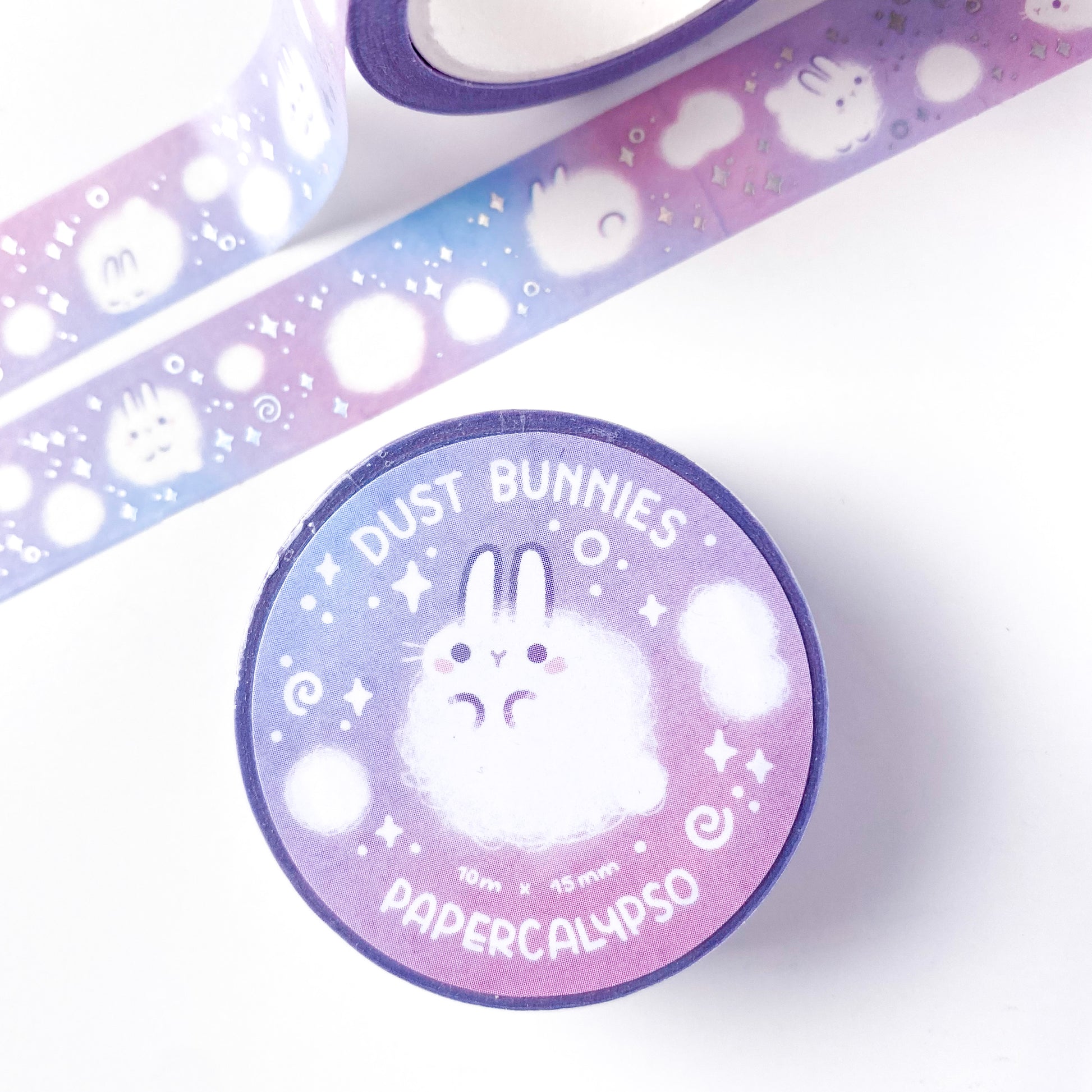 The roll of washi tape is packaged with shrink wrap plastic and a round sticker on one side. On it is an adorable fluffy bunny looking at us. It reads "Dust Bunnies, 10 metres by 15 milimetres, by Papercalypso" 