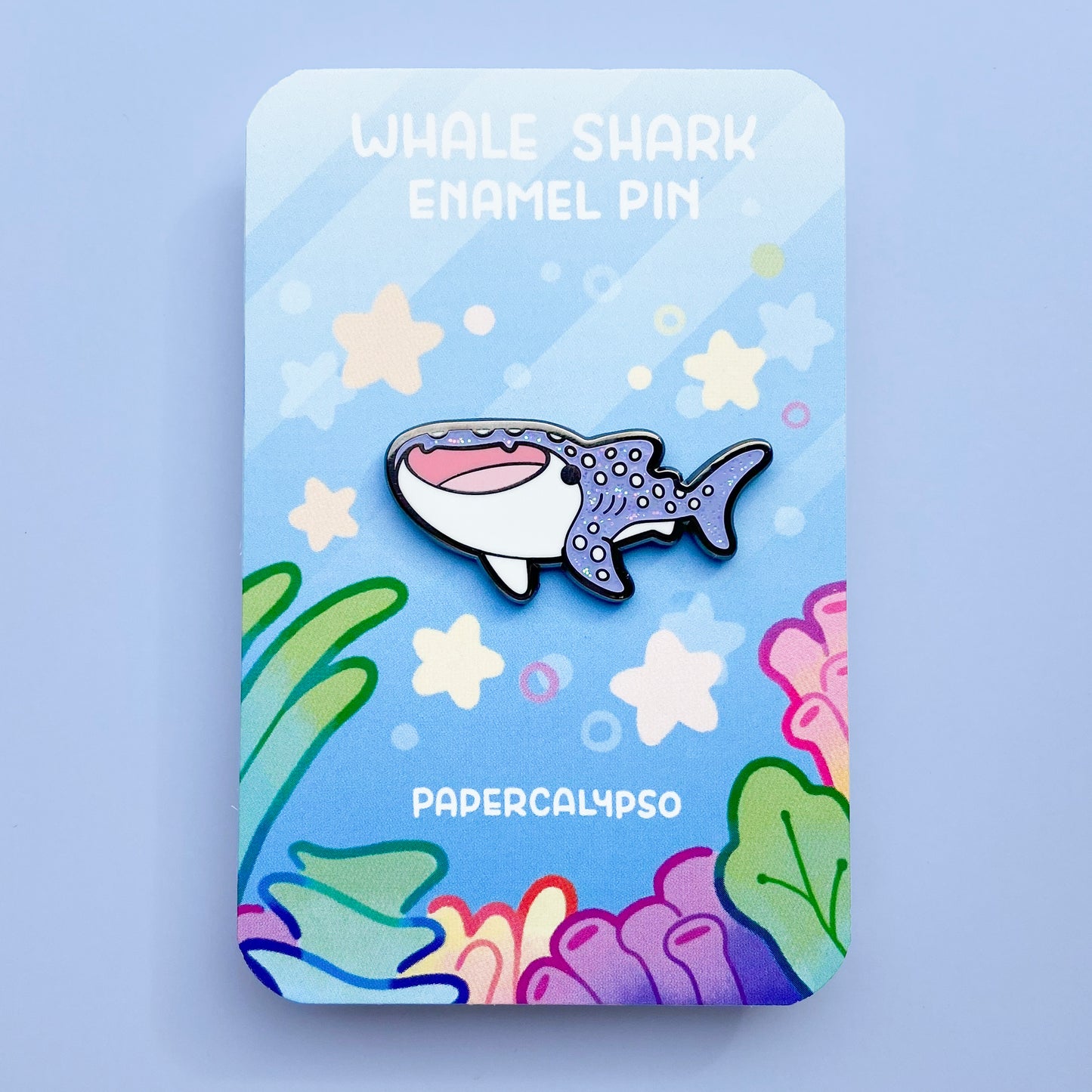 A black nickel hard enamel pin of a whale shark. He has a white tummy and glittery purple back with white spots. He's smiling with his mouth open and looks very cute! 